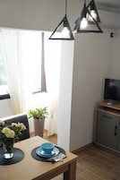 Uma sala de jantar com mesa posta para dois, um vaso de planta e uma televisão sobre um móvel.