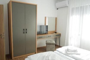 Quarto com guarda-roupa, escrivaninha com espelho, televisão e cama com roupa de cama branca impecável.