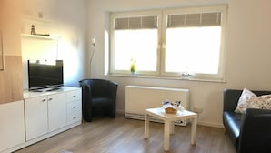 A modern living room with a flat-screen TV on a white cabinet, a black leather chair, a white coffee table with a basket, and a black sofa with floral cushions. - House Nicole vacation apartment 1 (Cuxhaven)