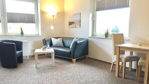 A living room with a dark leather sofa, a white coffee table, a small table with a glass, and a window with blinds. - House Nicole vacation apartment 3 (Cuxhaven)