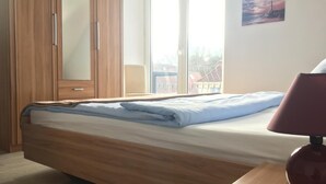 A bedroom with a wooden bed, a desk with a lamp, a wardrobe, and a window with a view of buildings. - House Nicole vacation apartment 5 (Cuxhaven)