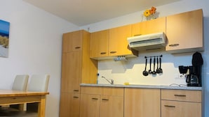 Een kleine keuken met houten kastjes, een wit aanrechtblad en een eethoek met een tafel en stoelen.