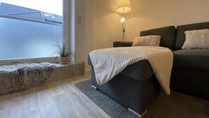 Un salon moderne avec un canapé foncé, un plaid blanc, une lampe sur pied et une vue sur un immeuble par le biais d’une grande fenêtre.