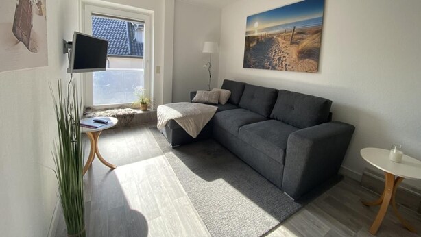 Un salon moderne avec un canapé gris, une petite table ronde, un téléviseur et un tableau représentant une scène de plage.