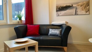 A living room with a black leather sofa, a wooden coffee table, a red curtain, a potted plant, and a beach-themed painting on the wall. - House Nicole vacation apartment 10 (Cuxhaven)