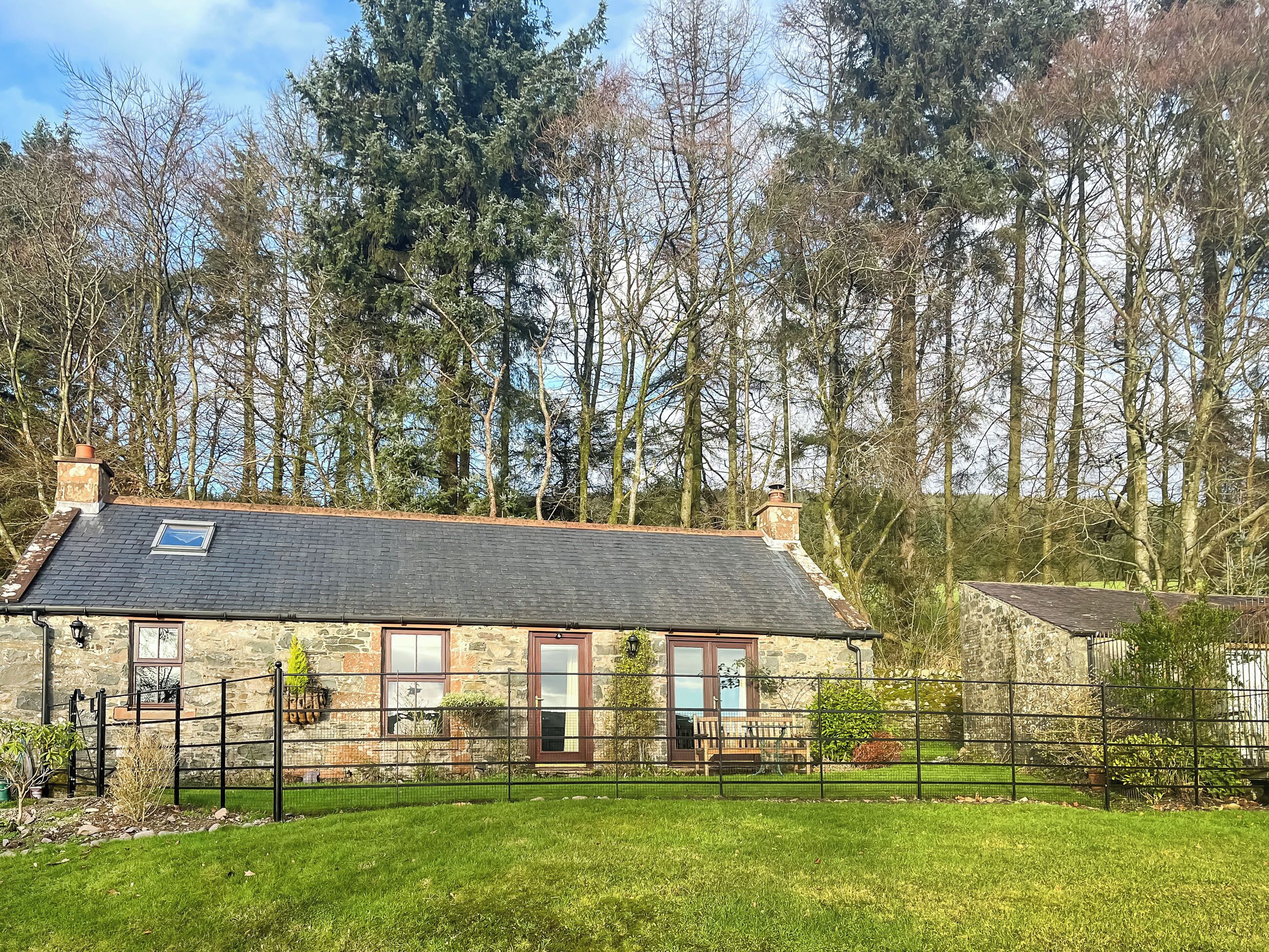 Une maison en pierre avec un toit en tuiles, une cour clôturée et une vue sur une forêt.