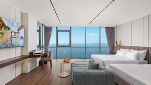 Habitación de hotel moderna con un ventanal grande que ofrece vistas al mar, un sofá, una cama, un escritorio y un televisor mostrando una imagen panorámica.