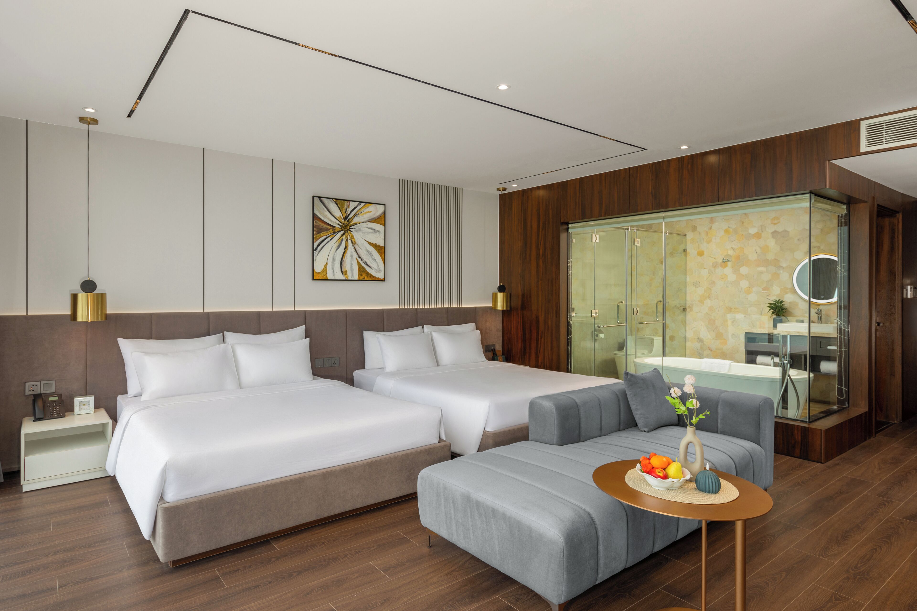 A modern hotel room with two beds, a sofa, a small table with fruit, and a bathroom with a glass shower enclosure.