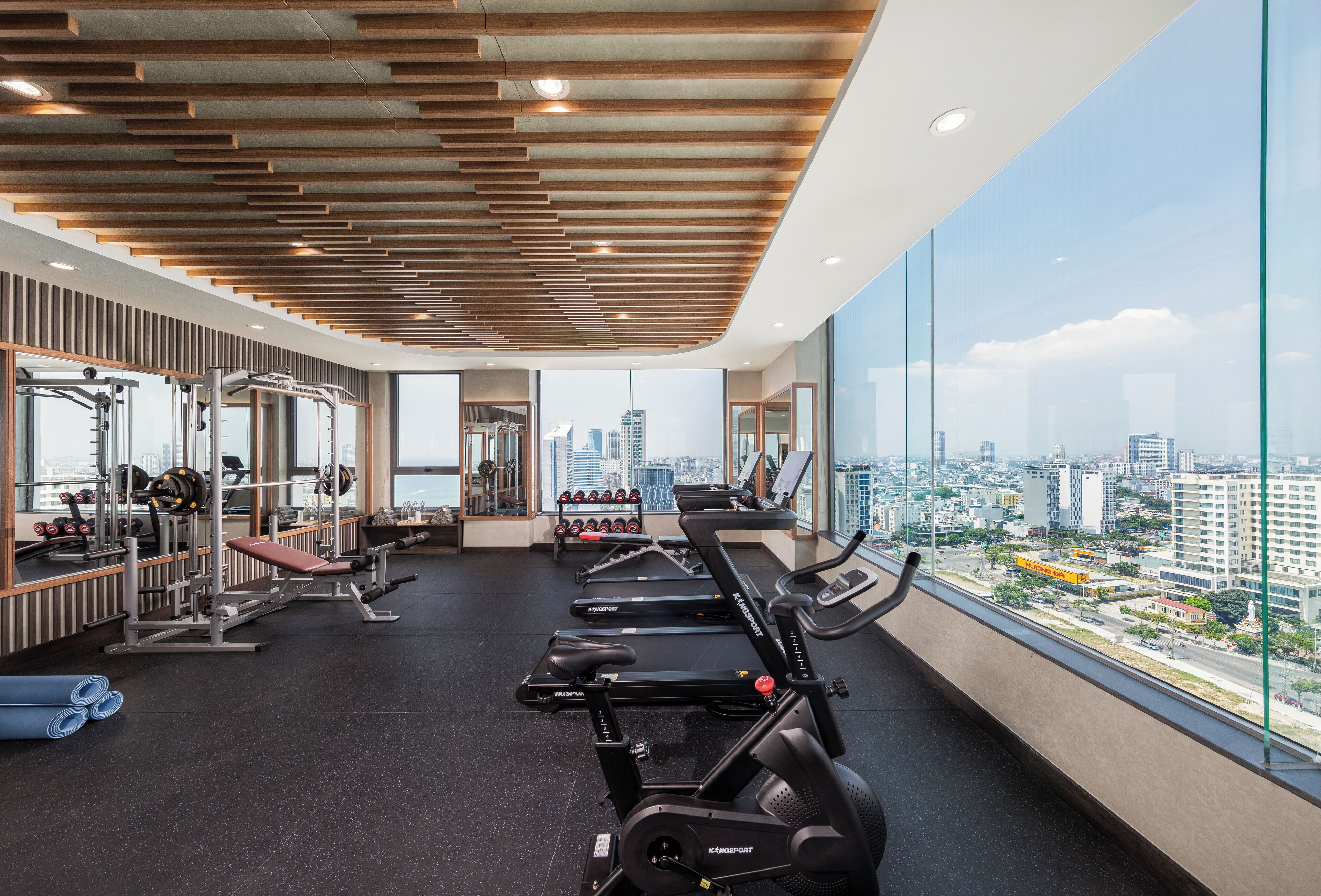 Uma academia com esteiras, halteres e aparelhos de musculação, grandes janelas com vista para a cidade.
