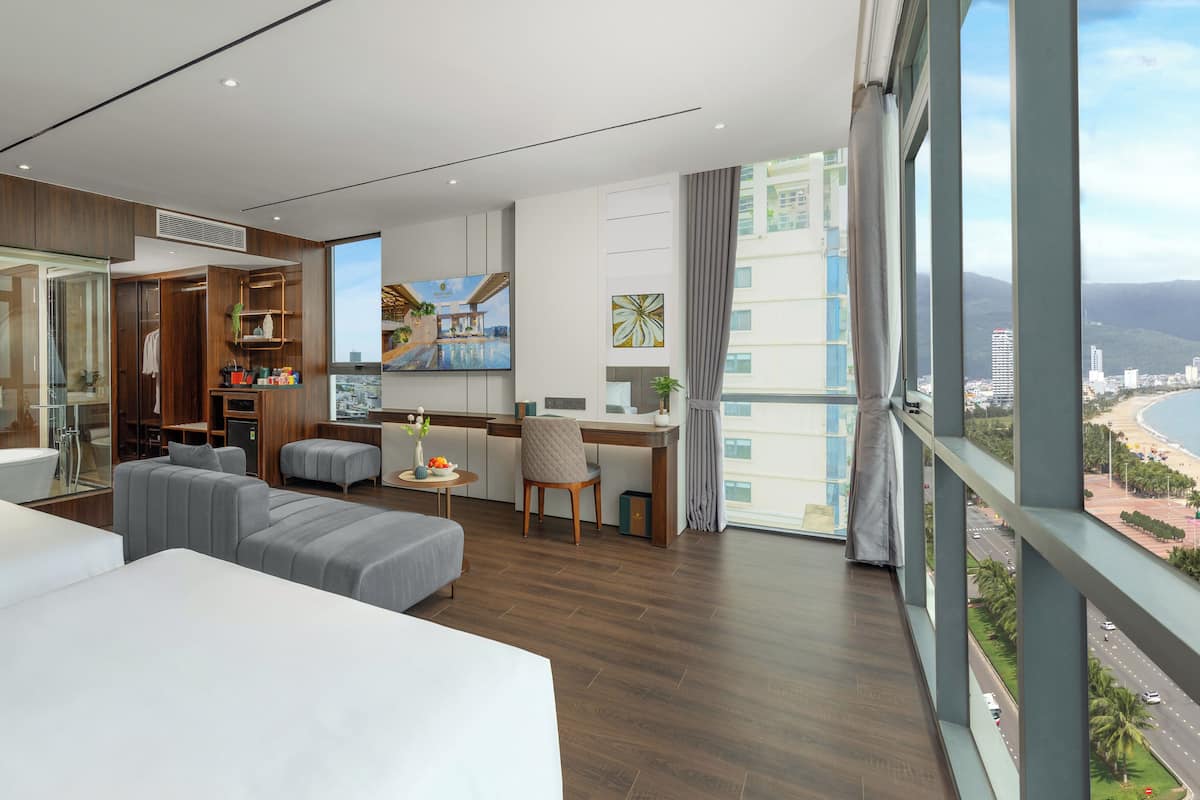 A modern hotel room with a large bed, a desk, a sofa, and a view of the city and beach.