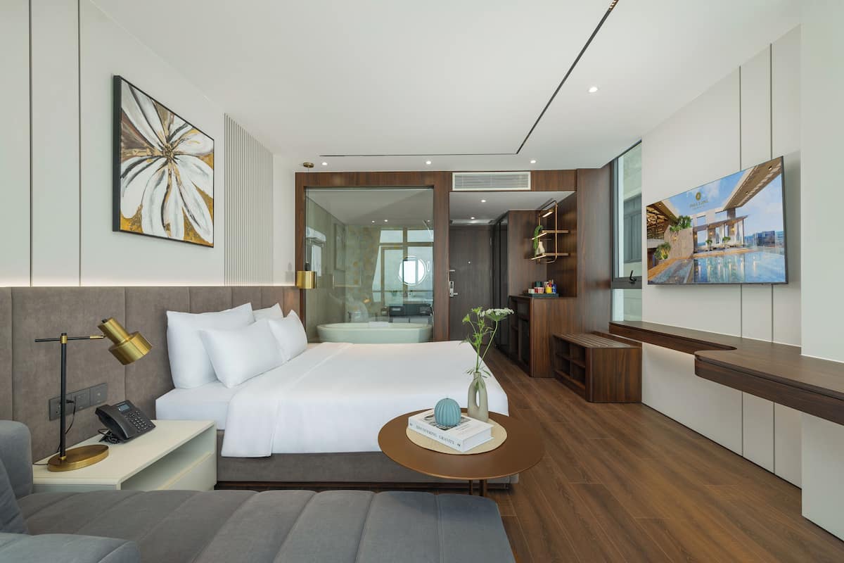 A modern hotel room with a large bed, bedside table with a lamp, a small round table with a vase and book, and a large window offering a view of the pool area.