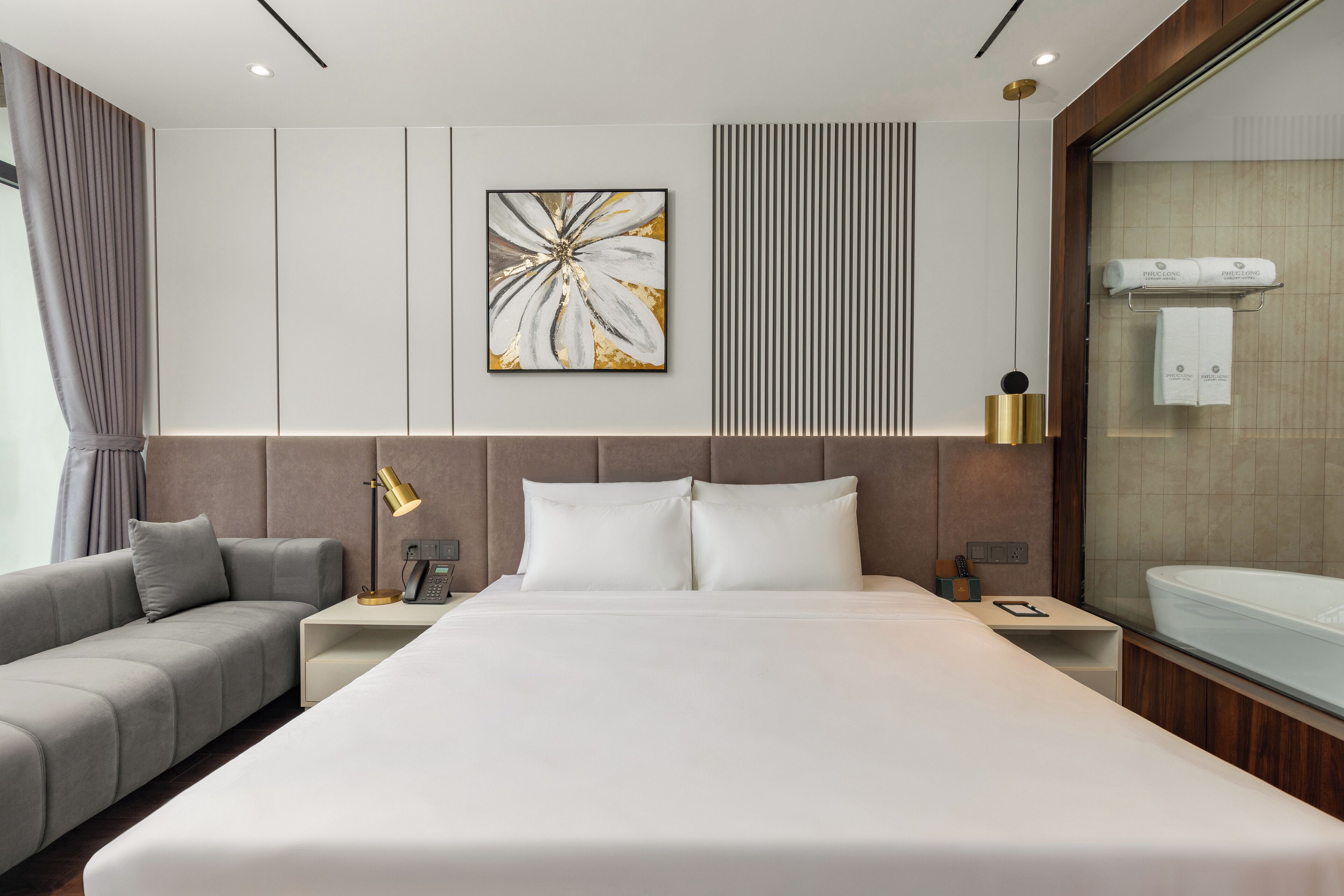 A modern hotel room with a large bed, a sofa, a bedside table with a lamp, and a wall-mounted towel rack.