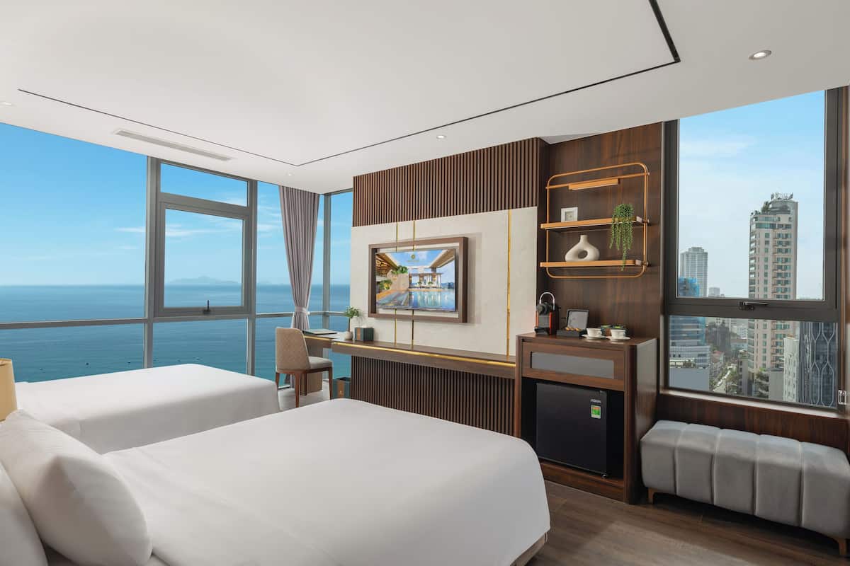 A modern hotel room with two beds, a TV, a desk, and a view of the ocean.
