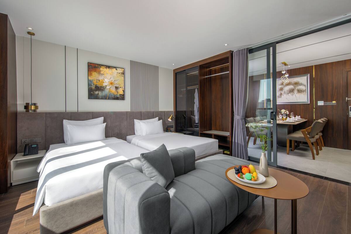 A modern hotel room with two beds, a sofa, a dining area, and a bathroom.