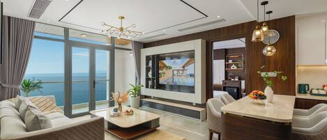 A modern living room with a large window, a sofa, a coffee table, and a dining area with a kitchen in the background.