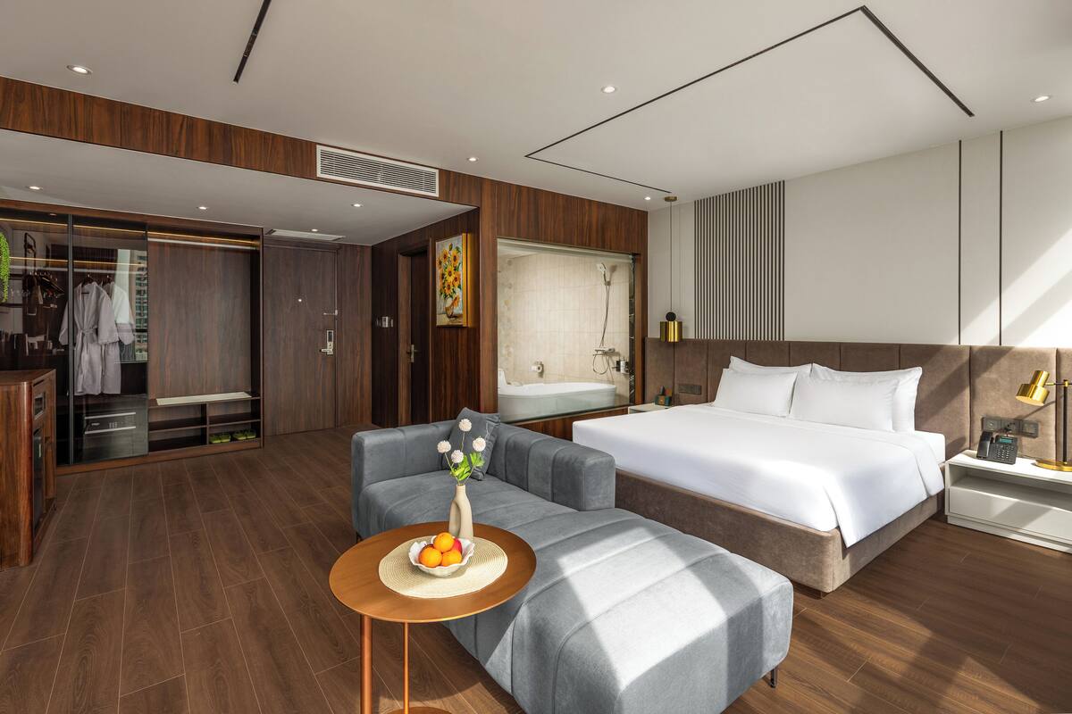 A modern hotel room with a large bed, a sofa, a small table with fruit, a walk-in closet, and a bathroom with a bathtub and shower.
