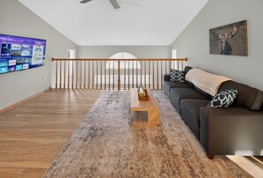 Un salón amplio con una mesa de centro de madera, un sofá gris y un televisor instalado que muestra varios canales.