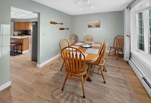 A dining area with a round table and six chairs, a kitchen in the background, and a window with curtains. - Maplewood Lake House - Boat Rentals, Private Dock (Newport)