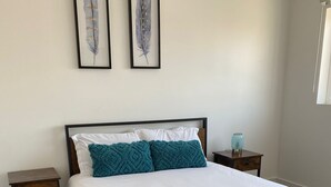 Una cama bien hecha con sábanas blancas y almohadas turquesa, una mesita de noche con una lámpara, y dos cuadros con plumas en la pared.