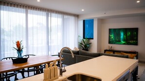 A modern kitchen with a built-in sink, a dining area with a table and chairs, a sofa, and a large TV screen displaying a forest scene. - Happy Hour by Experience Jervis Bay (Huskisson)