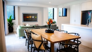 A modern kitchen with white cabinets, a wooden dining table, and chairs. There is a sofa and a potted plant in the living area. - Happy Hour by Experience Jervis Bay (Huskisson)