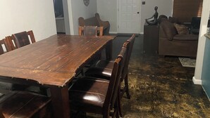 Een eetgedeelte met een houten tafel en stoelen, een witte deur en een bruine leren bank op de achtergrond.