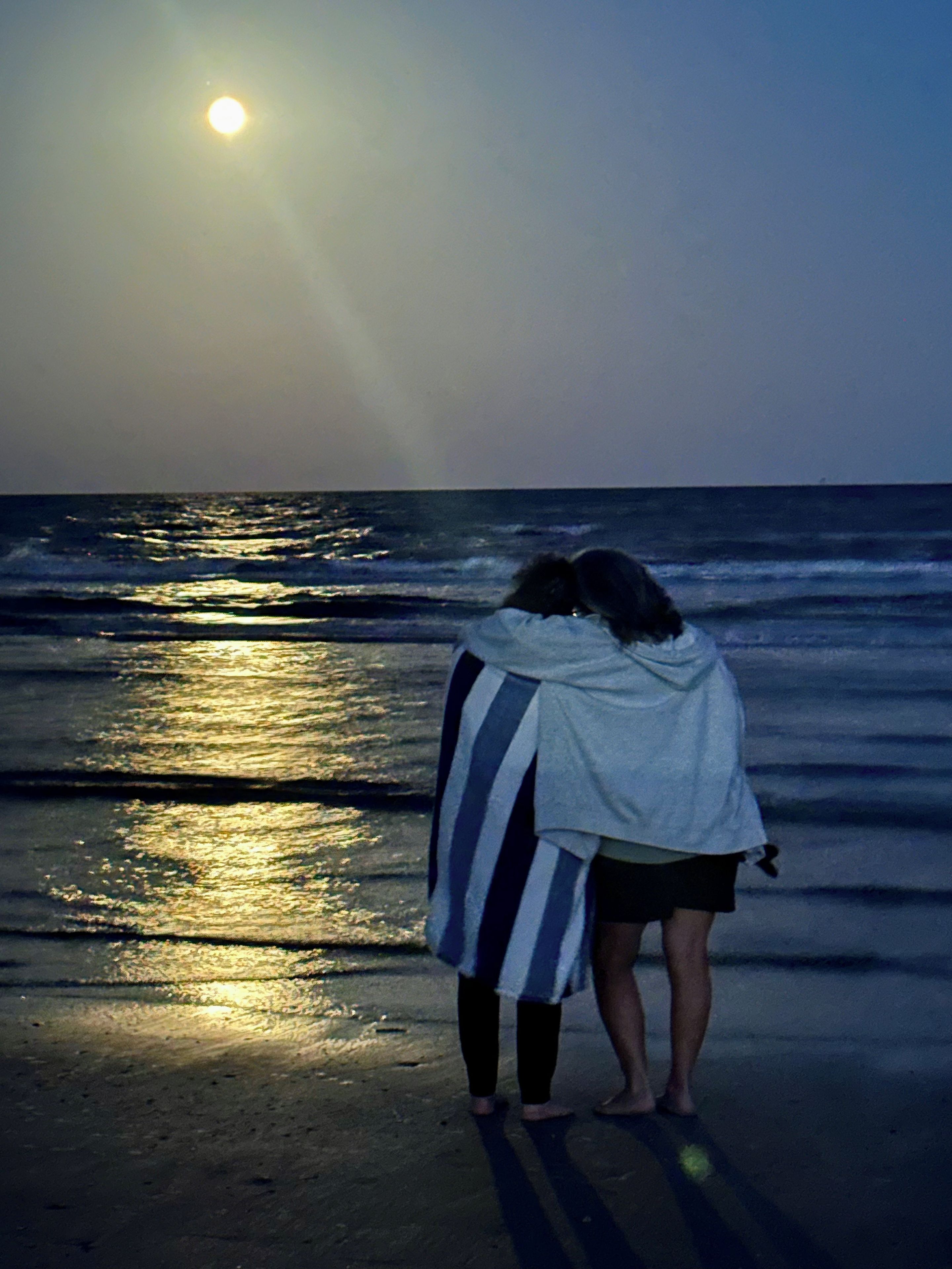 A beach scene with a single person standing on the sand facing the ocean, with the sun low on the horizon.