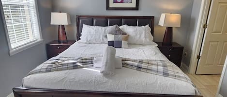Un dormitorio con una cama grande, dos mesitas de noche con lámparas, un cuadro en la pared y una ventana con persianas.