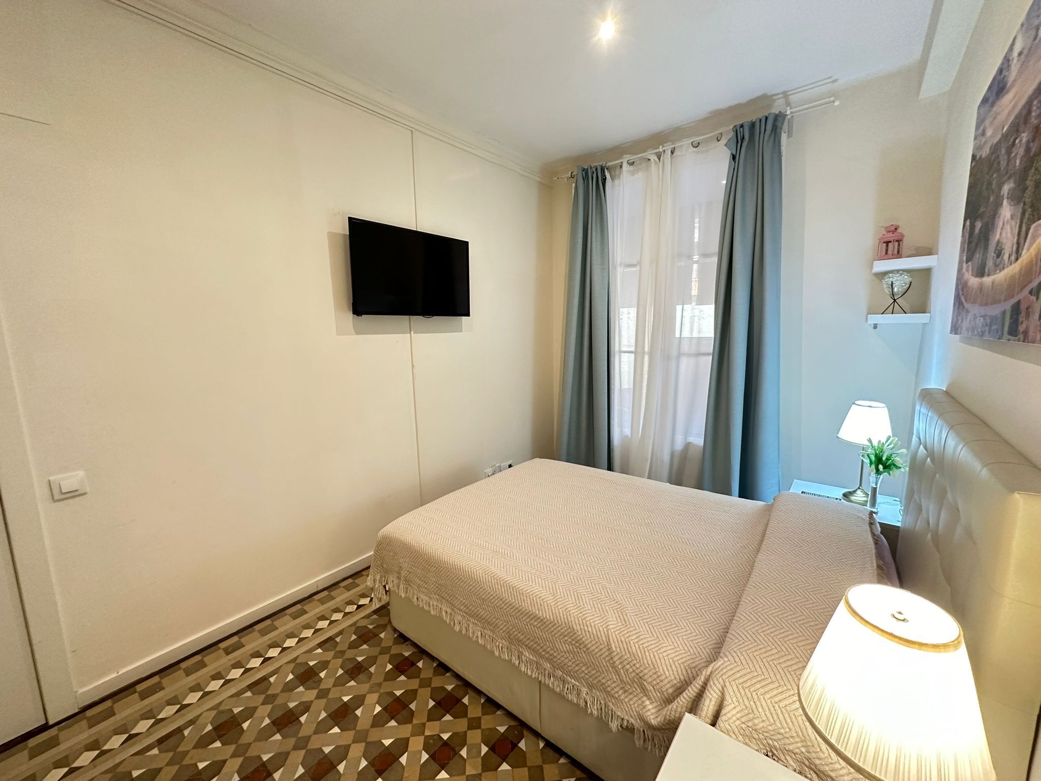 A bedroom with a bed, a TV mounted on the wall, a lamp, and a window with curtains.