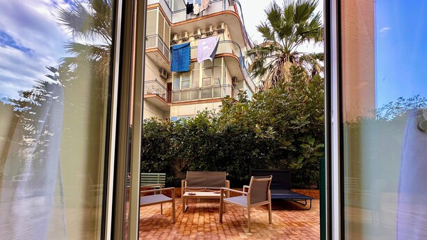 Vista attraverso una porta di vetro su un cortile con pavimento in mattoni, arredi da esterno e un edificio con balconi sullo sfondo.