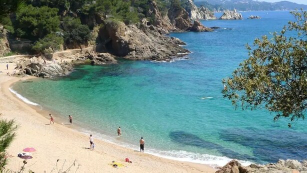 Una playa con agua turquesa cristalina, acantilados rocosos y algunas personas disfrutando del sol.
