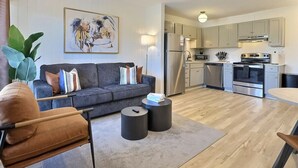 Ein modernes Wohnzimmer mit grauem Sofa, einem Couchtisch und einem Essbereich mit Tisch und Stühlen. Die Küche ist sichtbar, mit Edelstahlgeräten und hellen Schränken.