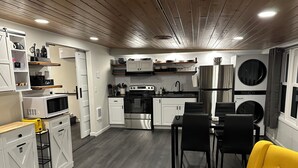 Een compacte keuken met witte kasten, een roestvrijstalen koelkast en een eethoek met zwarte stoelen en een gele bank.