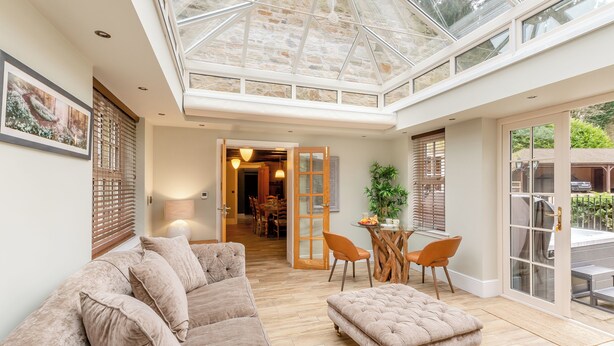 Une pièce spacieuse avec un plafond haut, un toit en verre, un coin salon confortable avec un canapé et un pouf, une table à manger avec des chaises, et une vue sur l’extérieur.