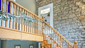 Un escalier en bois, avec un mur de pierres en arrière-plan, une porte donnant sur une pièce dont la porte est blanche, et deux tableaux encadrés accrochés au mur.