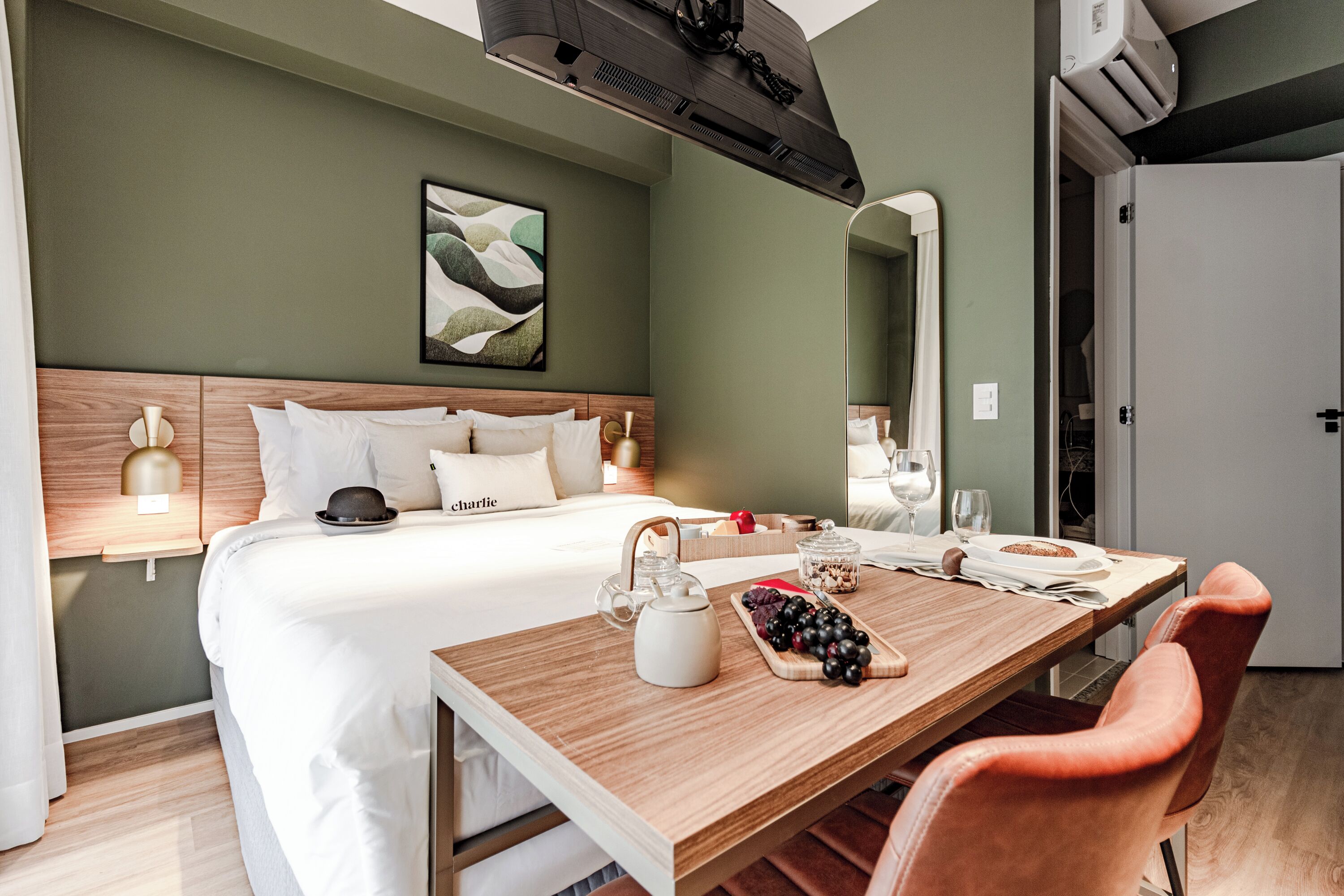 A modern hotel room with a neatly made bed, a wooden dining table with a teapot and grapes, and a large mirror on the wall.