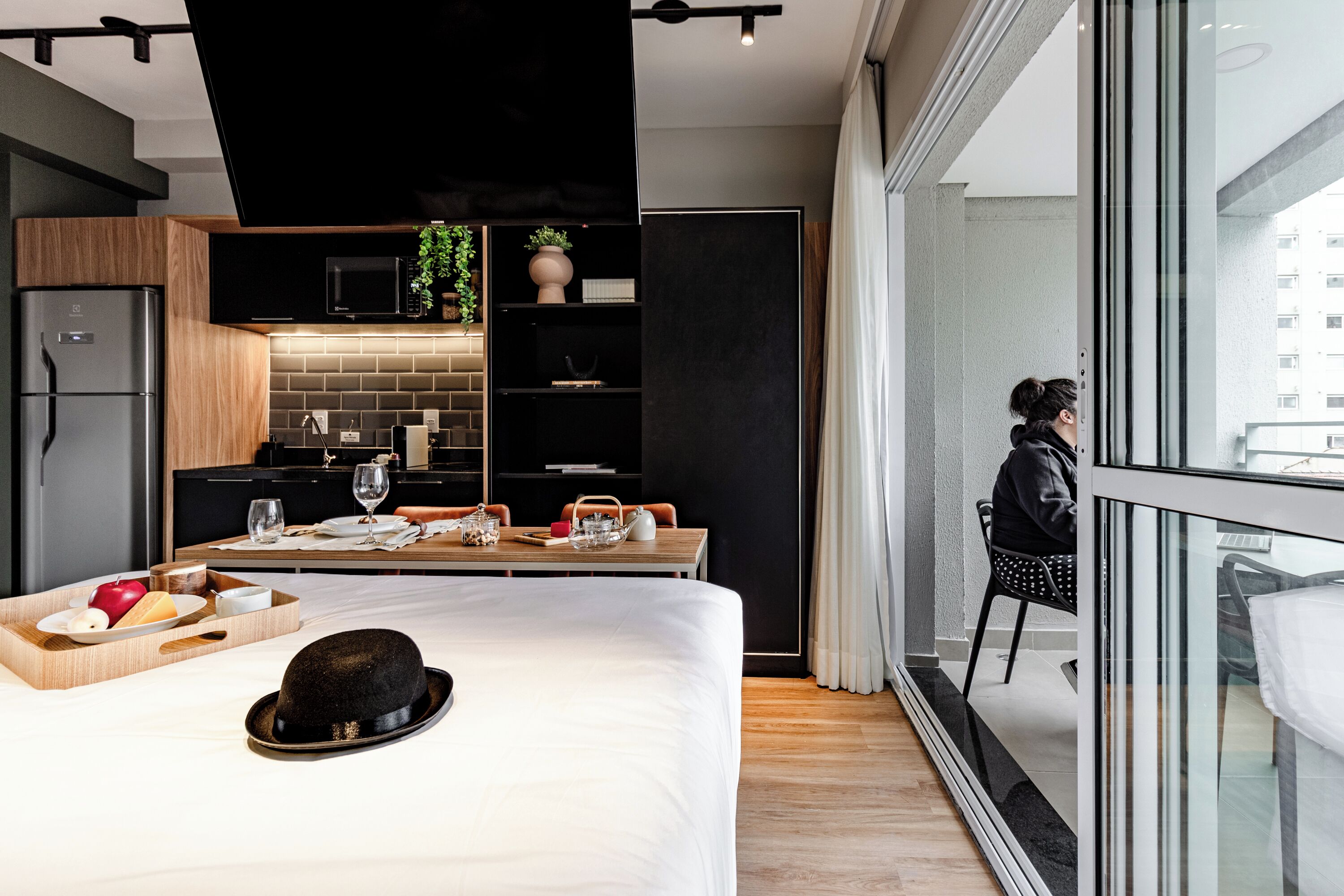 Quarto de hotel moderno com uma cama grande, área de refeições, cozinha e vista da cidade através de uma janela ampla.