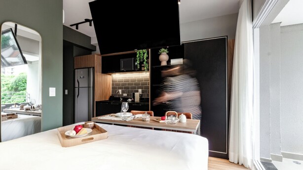 Une chambre d’hôtel moderne dotée d’un grand lit, d’un coin repas avec une table dressée pour deux personnes, d’une cuisine équipée d’un four à micro-ondes et d’un évier, et d’une porte coulissante donnant sur un balcon.