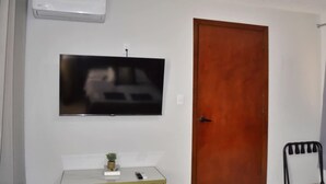 A room with a mounted TV, a wooden door, a small table with a plant, and a chair. - Céntrico y Confortable Departamento 101 (San Luis Potosí)