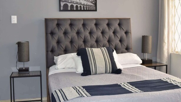 A bedroom with a tufted headboard, a bed with white and patterned bedding, a framed picture above the bed, and two bedside lamps. - Céntrico y Confortable Departamento 101 (San Luis Potosí)