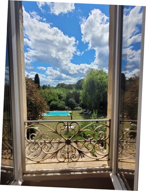 Vista da janela com grades de ferro ornamentadas, céu azul límpido e paisagem verdejante com árvores e piscina.