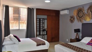 Hotellihuone, jossa on kaksi sänkyä, ikkuna verhoilla, seinään asennettu ilmastointilaite ja koristeellinen seinäelementti.