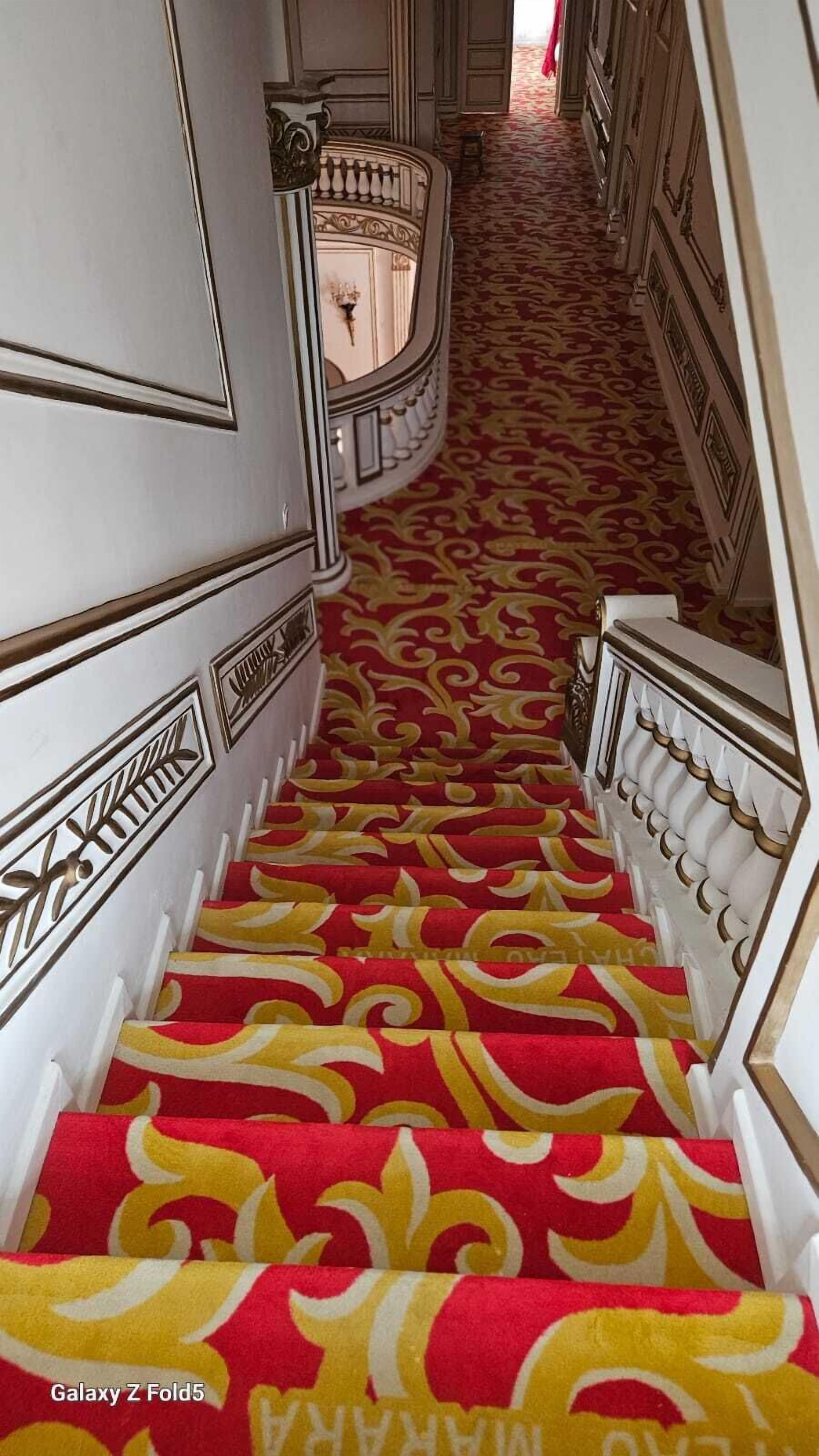 A grand staircase with a red and yellow patterned carpet, white walls, and ornate balustrades.