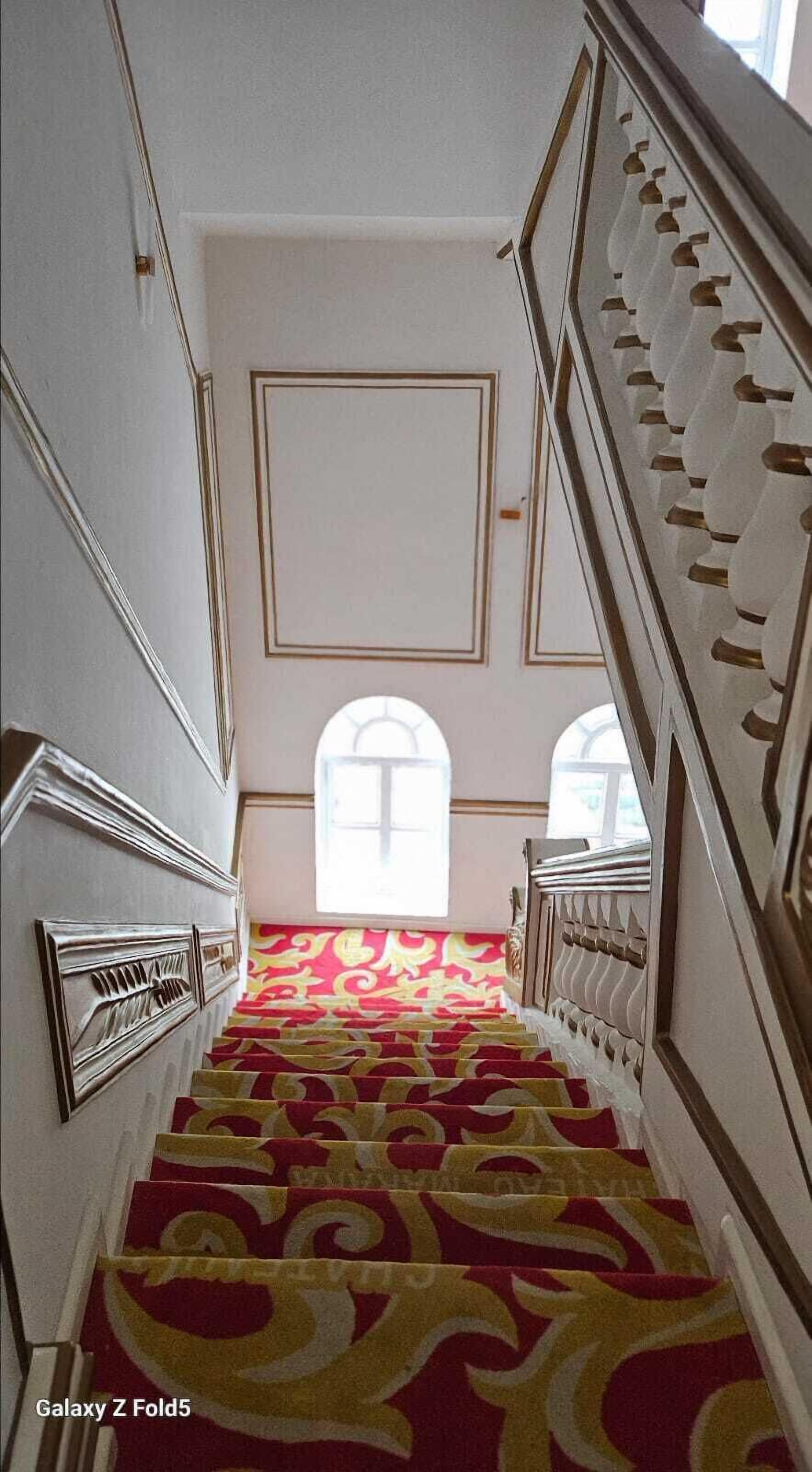 Una scala con un tappeto decorato, pareti bianche e una grande finestra in cima.