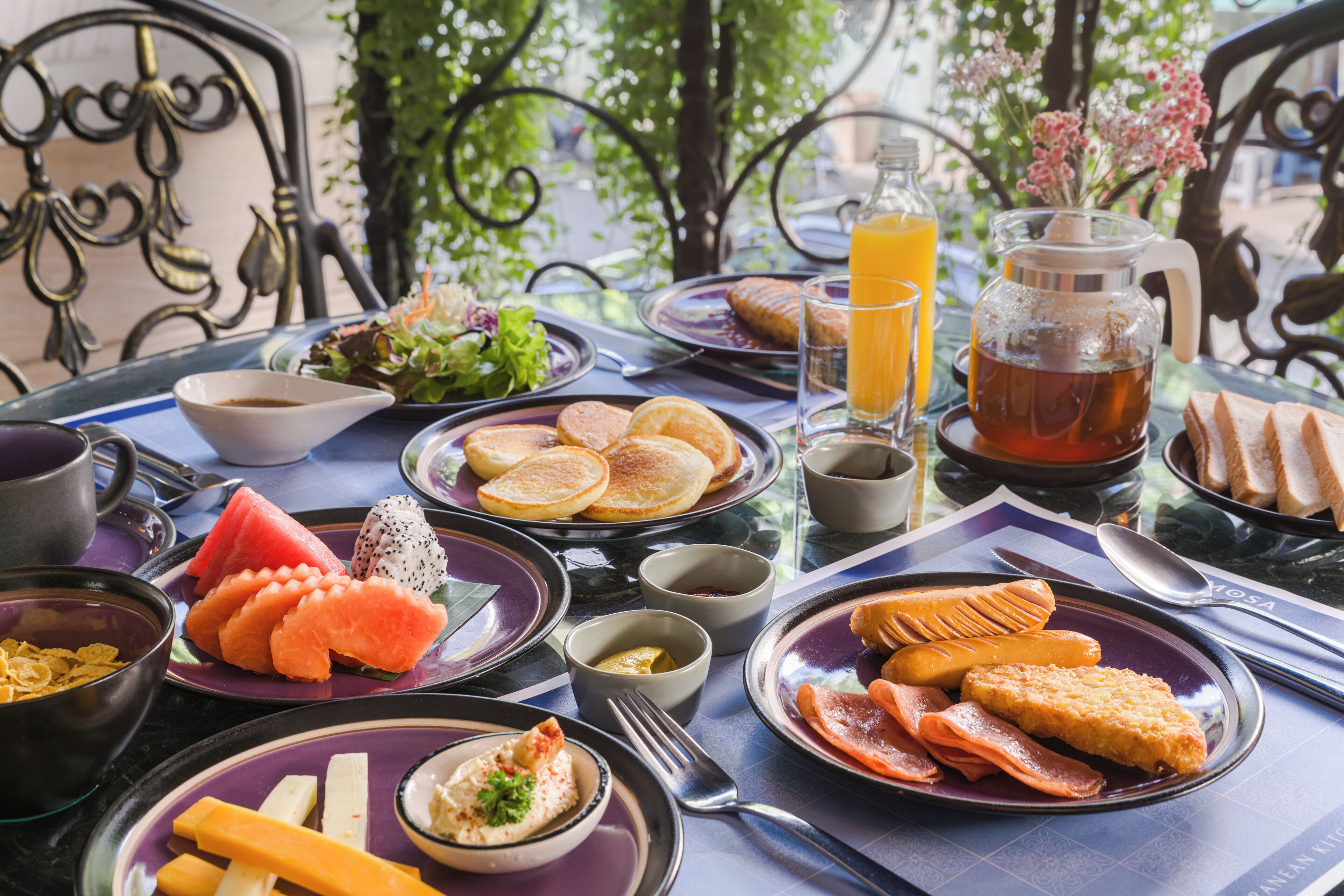 Un desayuno con diversos platos, como panqueques, huevos y fruta fresca, sobre una mesa con vista a follaje.