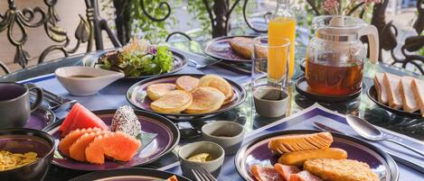 Un desayuno con diversos platos, como panqueques, huevos y fruta fresca, sobre una mesa con vista a follaje.