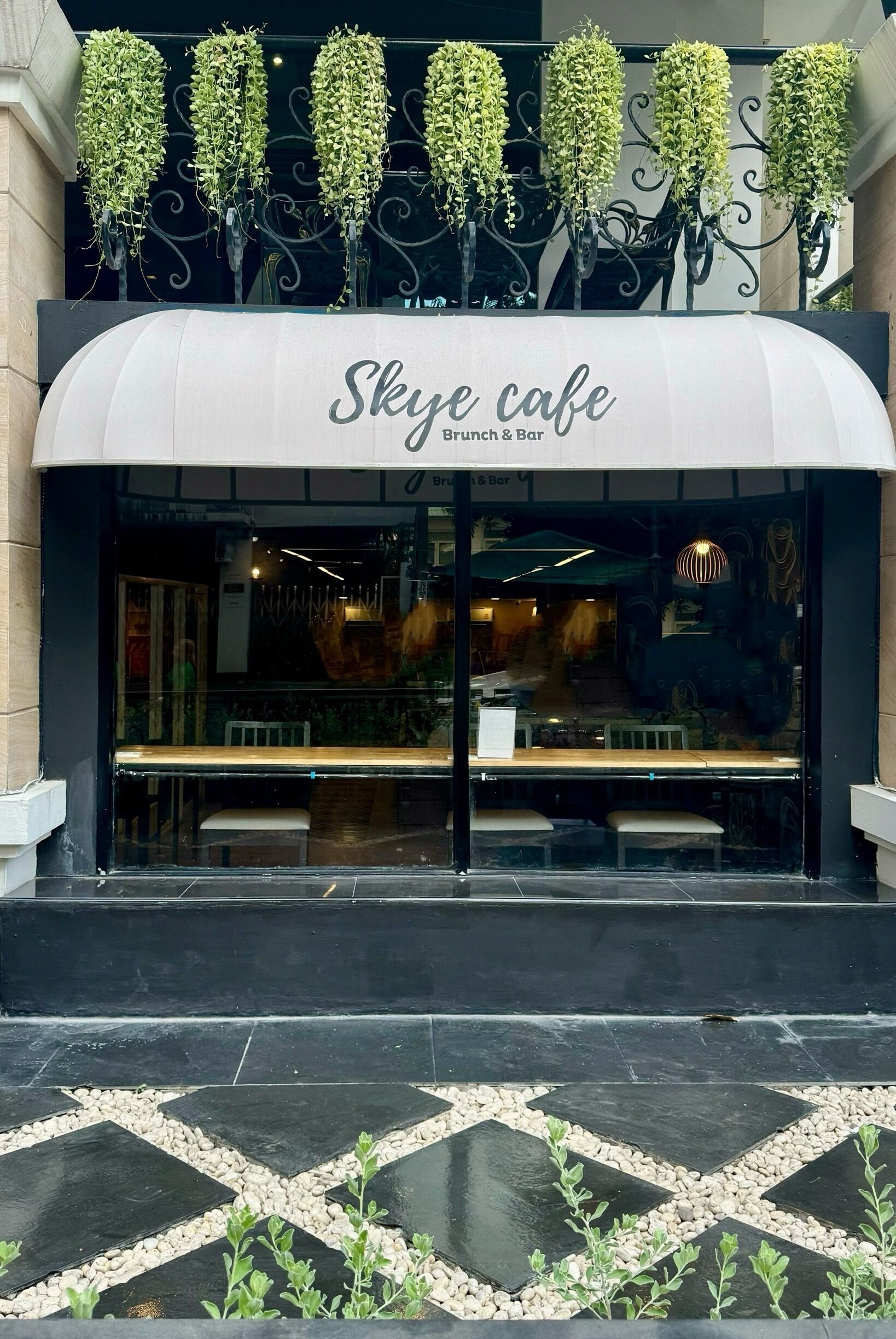 Exterior del Sky Cafe, con un diseño moderno que incluye un toldo curvo, zona de estar interior y plantas colgantes decorativas.
