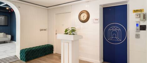 Une salle d’attente avec une porte bleue, un comptoir d’accueil blanc, un banc vert et une horloge au mur.
