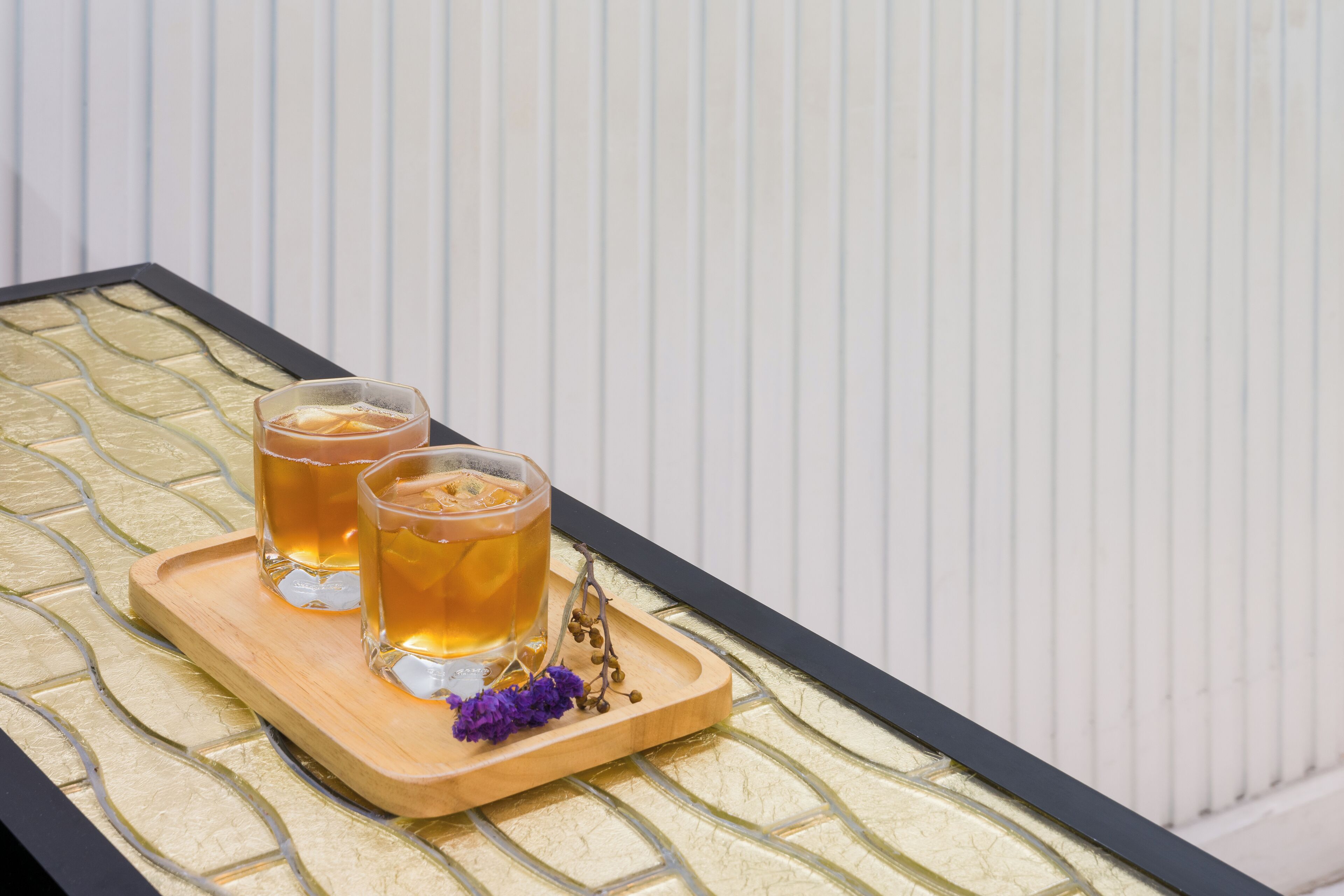 Dos vasos de líquido ámbar sobre una bandeja de madera con una rama decorativa, colocados sobre una mesa de vidrio texturizado frente a una pared blanca con paneles.