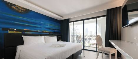 A modern hotel room with a large bed, a desk, and a view of the city through large windows.